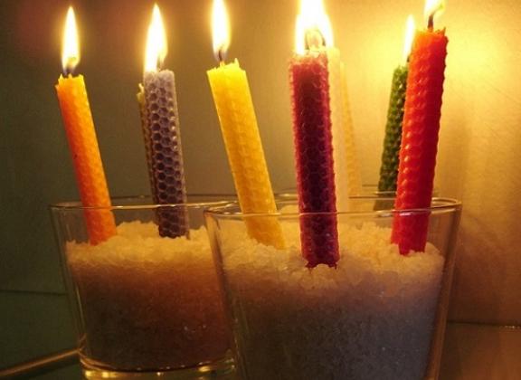 Kuvassa erivärisiä mehiläisvahakynttilöitä "istutettuina" kirkkaisiin laseihin. Kynttilöissä palaa tuli. 